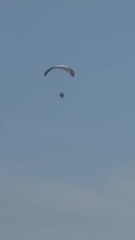 This video shows a person paragliding over a body of water. The paraglider appears to be a solo pilot, and the paraglider wing is a light color with some red accents. The person is suspended from the wing by a harness and a small motor is attached to the harness to provide propulsion. The paraglider is flying at a relatively low altitude, and the water below is calm and clear. There are some small waves breaking on the shore, and a beach with several umbrellas and people can be seen in the distance. The sky is a clear blue with a few wispy clouds. The video appears to have been filmed from a high vantage point, possibly from a hill or a building overlooking the beach and the water. The paraglider is moving from left to right across the frame. There are no other visible aircraft or boats in the immediate vicinity. The overall impression is one of peaceful recreation and a beautiful day for flying.