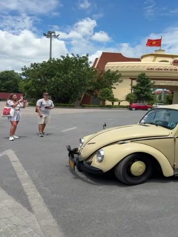 The video pans across a beige Volkswagen Beetle parked on an asphalt surface, likely a parking lot or road. Two people, a woman and a man, stand to the left of the car, seemingly posing for photos. The woman, in a striped dress, holds up a phone to take a picture. The man, in a t-shirt and shorts, also holds a phone.

Behind the people and the car, several large red and brown trucks are parked, suggesting a commercial or transport hub. The background includes trees, a large building with a red roof, and a cloudy blue sky, indicating it is daytime with mixed weather. The car itself has several stickers on its doors and rear window. One prominent circular sticker on the driver's side door reads "LAOS VOLKSWAGEN CARAVAN 07" with text below it. The license plate is visible and reads "DZ 1724".

The overall scene appears to be part of a car rally or a special event involving Volkswagen vehicles, given the "Laos Volkswagen Caravan" sticker. The people's demeanor suggests a casual and perhaps excited mood, typical of participating in such an event. The setting is outdoors in what seems to be a warm climate, indicated by the palm trees and attire of the people. The presence of trucks and the building structure suggests a location in a town or near a port, potentially an entry/exit point for transport. The time of day is clearly during daylight hours.