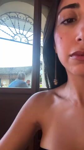 The video begins with a close-up shot of a woman's face, partially obscured. The camera then pans out to reveal an outdoor seating area with tables and chairs covered in white tablecloths. Beyond the seating area, a scenic view of buildings, trees, and a tower can be seen under a cloudy sky. A man is visible in the reflection of a window, facing away from the camera. The overall scene suggests an outdoor dining setting with a picturesque view.