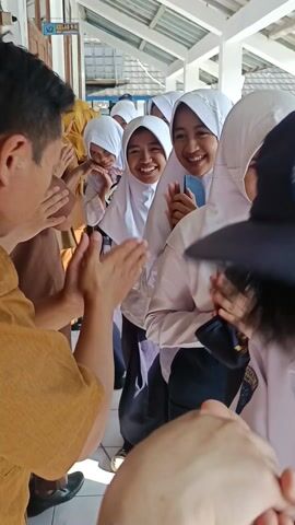 A group of smiling young women in white hijabs walk through a line of adults, some wearing yellow clothing, who are clapping for them. The girls hold their hands in a prayer-like gesture, and one of them carries a dark blue cap. They are in a brightly lit, covered walkway, with the words "KELAS 9-H" visible on a sign. Tiles make up the walkway floor.