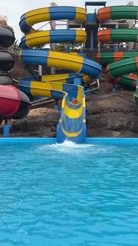 A person in a red swimsuit slides down a blue and yellow water slide, splashing into a clear blue swimming pool. The water park setting features multiple large, enclosed water slides in various colors, including blue and yellow, red and black, and orange and green, all winding down a rocky, earthen mound. The skies appear overcast, indicating daytime. Faint text reading "AQUA" is visible on a structure in the upper left background. The scene captures a moment of fun and activity at the water park, located in La Oliva, Spain.