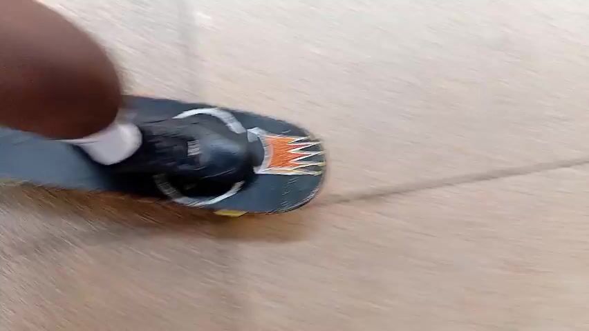 A first-person perspective video shows a skateboarder's feet and the skateboard moving across a light-colored, linoleum-like floor with faint grid lines. The skateboard is dark with orange and white markings. The skateboarder's feet are visible, one in a dark shoe and the other in a white sock, with a black knee pad on. The motion blur indicates movement, suggesting the skateboarder is riding. The environment appears to be indoors, possibly a school or a public building due to the flooring.