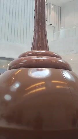 The video shows a chocolate fountain installation in a modern building. The fountain has a large base, followed by a tiered section, and then a tall, slender column of flowing chocolate that reaches to the ceiling. The ceiling is a glass dome with a circular design, allowing natural light to filter in. The building's interior features concrete walls with vertical ribbed patterns and large windows with curtains. The overall impression is one of contemporary design and a focus on the chocolate fountain as a central, artistic feature.