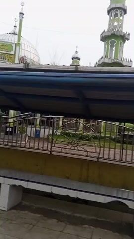 A green and white mosque minaret is visible in the background of the video. In the foreground, a metal gate with horizontal bars is partially open, revealing a building with an orange facade. Signs are visible on the building, one reading "RAUDHATUL ATHFAL NURUL AMALIYAH". There are some plants and debris in the lower part of the frame. The weather appears overcast.