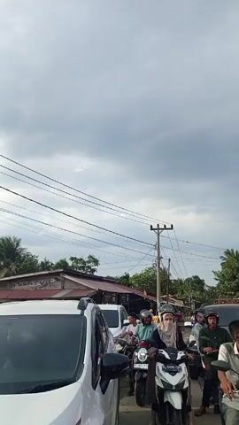 The video shows a congested street scene in what appears to be Southeast Asia, likely Indonesia given the license plates on the vehicles. The sky is overcast, suggesting a cloudy day. The foreground features cars and numerous scooters/motorcycles, all stopped in traffic. Many riders are wearing helmets. Some women are wearing hijabs. In the background is a construction zone, marked by a white and red barrier, with cranes visible. Signage indicates "Dilarang Masuk Area Konstruksi" which translates to "No Entry Construction Area." Overhead power lines crisscross the scene.