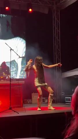 A woman with long, dark hair performs on a stage. She is wearing a light-colored, sleeveless dress with a white hem and white boots that reach her knees. She has a white wristband on her left wrist. Her arms are outstretched, and she appears to be in motion.

Behind her, a large screen displays a white, abstract image. To her left, a microphone stand is positioned on the stage. Further left, a person is visible behind a DJ setup, illuminated by red and blue lights. Red lights are also visible on the ceiling above the stage. Smoke or haze drifts across the stage. The scene suggests a live music performance or concert.

The stage itself is dark, with red lighting on the front edge. Metal trussing is visible on the right side of the frame. In the foreground, the blurred silhouette of an audience member's head and shoulders can be seen. The lighting and the presence of a stage setup indicate that the event is taking place in the evening or at night.