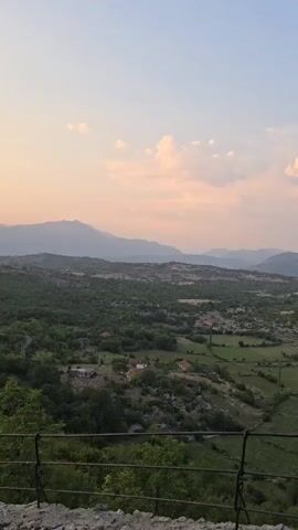 A high-angle, slow pan shot shows a rural landscape at dusk. The sky is a gradient of pale blue to pink with scattered clouds. Rolling hills covered in green foliage and trees stretch across the middle ground, dotted with small houses with red roofs. In the foreground, a dark metal railing runs along the bottom of the frame. The overall atmosphere is peaceful and serene.