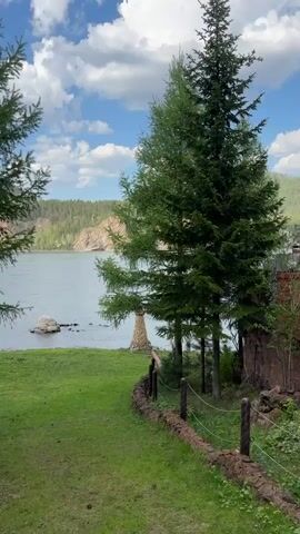The video shows a scenic view of a river landscape. In the foreground, there's a green grassy area with a low stone wall and a rope fence. Several evergreen trees are visible, obscuring some of the view.  A small stone structure, resembling a miniature tower or cairn, sits near the water's edge. Beyond the trees and grass, the river stretches out, dotted with a few rocks.  Across the river, there is a rocky cliff partially covered with trees and other vegetation. The sky is blue with scattered white clouds.  The video appears to be taken during a calm and pleasant day.