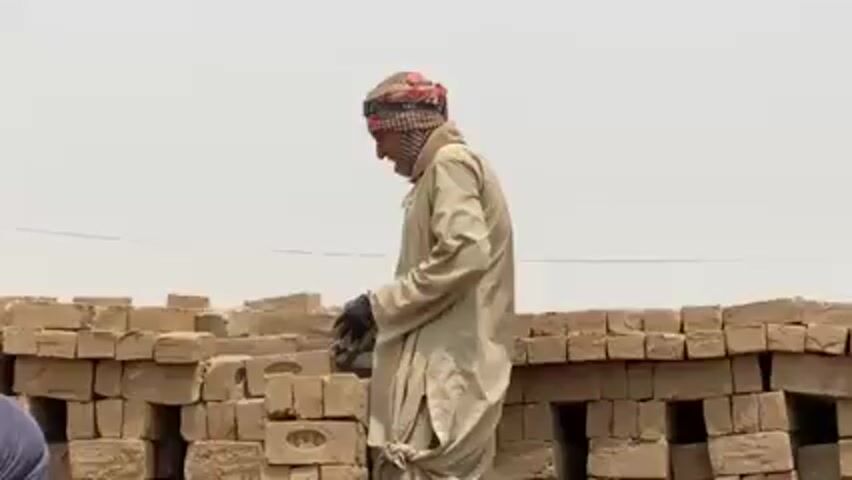 A man wearing a tan robe and a head covering is standing next to a large pile of light brown bricks. He is wearing black gloves and appears to be inspecting or handling one of the bricks. The sky is overcast. The overall impression is a simple scene of labor or construction in a potentially hot and dusty environment.