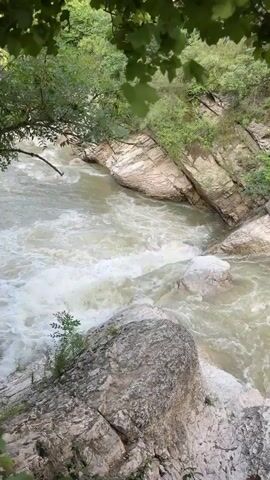 A fast-flowing river crashes over rocks in a narrow gorge. Steep, rocky cliffs line the sides of the gorge, covered in green foliage. Large rocks are scattered in the riverbed. Trees with green leaves hang over the top of the scene, casting shadows.