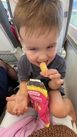 A young child sits on a train, holding a snack and eating it. The child is wearing a dark gray t-shirt and has light brown hair. They are holding a yellow, puffed snack in their right hand and bringing it to their mouth. In their left hand, they hold an open bag of snacks with pink packaging. The bag has a label that reads "SMILEAT" and "minitos". The child is seated on a light-colored seat, and the background shows the interior of a train with other seats visible. The child's legs are resting on a pink fabric, and a brown woven object is partially visible in the lower right corner. The child's expression is one of enjoyment.