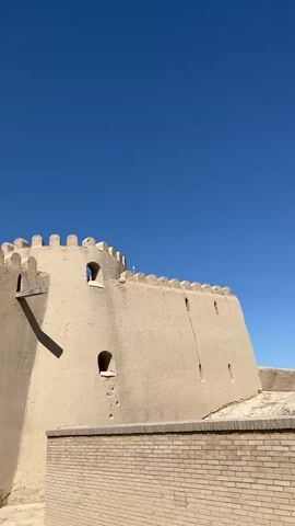 The video shows a section of an ancient mud-brick fortress under a clear blue sky. The fortress wall is tan and features crenellations along the top, with small, rectangular openings dotted along its height. A rounded tower-like structure is visible on the left side of the frame, casting a long shadow. In the foreground, a lower wall made of light-colored bricks is visible. The entrance to the fortress, a large arched gateway, is partially visible on the left. Through the gateway, a white tent and some figures can be seen. To the right of the entrance, a bundle of dry reeds stands upright. A person in a dark shirt walks away from the camera towards the gateway. The overall environment appears to be a historical site or tourist attraction, possibly in a desert or arid region given the material of the fortress.