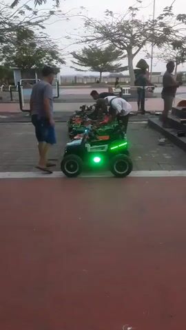 The video shows a row of small, green-lit electric ATVs parked on a paved area, possibly a boardwalk or park. Several men are interacting with the ATVs. One man in a gray t-shirt and blue shorts stands to the left, looking towards the ATVs. To the right of the ATVs, three men are visible. One man, wearing a white shirt and black pants, appears to be adjusting or preparing an ATV. Another man in a black shirt and jeans is leaning over an ATV. A third man in a gray shirt and shorts stands further to the right, looking at his phone. In the background, there are trees, a body of water, and exercise equipment. The lighting suggests it is either early morning or late afternoon.