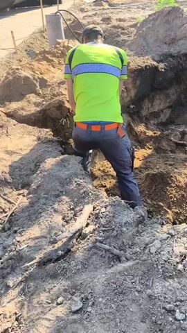 A person wearing a neon green shirt and gray pants is digging a hole in the dirt. The person is using a shovel and a power tool. The environment appears to be a construction site.