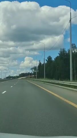 The video shows a view from a vehicle traveling on a highway. The road is paved and has white dashed lines marking the lanes, with a yellow line on the right side. There are streetlights lining the highway. On the left side of the road, there is a metal guardrail and trees. On the right side, there are trees and some construction or development visible in the distance. The sky is blue with large, fluffy white clouds. The overall impression is of a clear day on an open road.