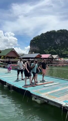 A group of six people are walking single file on a wooden pier. The pier is painted with blue and white stripes and is elevated above green water. In the background, there are several stilted buildings with red, orange, and brown roofs, suggesting a village on the water. A large, verdant, rocky island with steep cliffs rises dramatically behind the village. The sky is blue with scattered white clouds. The people are casually dressed, indicating a tourist setting. The person in the foreground is wearing a black dress and flip-flops, carrying a backpack. Other individuals are wearing shirts, shorts, and dresses, also in casual attire. The group is moving from left to right in the frame. The lighting suggests it is daytime, likely sunny.