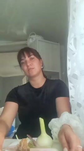 A woman with dark hair and bangs, wearing a black t-shirt, holds a white plate of food in front of her. The plate contains what appears to be a tea bag on a skewer, a piece of bread, a light-colored bulbous vegetable with a green stalk, and several pieces of pale, elongated food items, possibly meat or vegetables. The woman looks at the camera, then at the food, then touches her mouth and nose, and smiles. Her hair is partially braided. The background appears to be a kitchen, with a washing machine and cabinets visible. The lighting is bright, coming from the right.