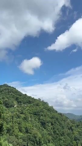 The video shows a mountain landscape under a blue sky with scattered white clouds. Lush green hills covered with dense forest stretch into the distance. The camera pans across the landscape, revealing more of the forested hills and the sky above.