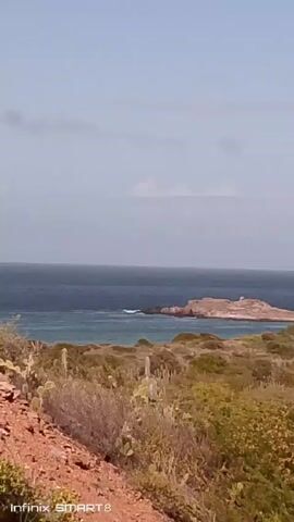 The video pans from left to right, showing a coastal landscape. At first, there's an island in the distance, surrounded by the ocean. In the foreground, there's dense, green vegetation with some cacti. As the camera pans, it reveals white buildings with red tile roofs, interspersed with palm trees. In the background, there's a large mountain. The sky is light blue with some white clouds. The video is labeled with "Infinix SMART 8" at the bottom.