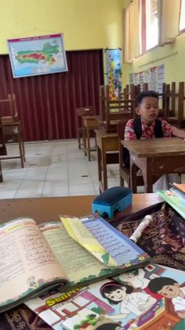The video opens on an open book with colorful illustrations on a desk in what appears to be a classroom. The book is partially obscured by other materials, including a patterned cloth and what looks like a pencil. The camera then pans to reveal a young boy, dressed in a traditional garment and a vest, sitting at a wooden desk. He appears to be speaking, his mouth open. Behind him, several empty wooden chairs are stacked on top of desks, suggesting the room is either preparing for class or is at the end of the day. A map of Indonesia is visible on the wall, along with other educational charts and posters. The lighting suggests an indoor environment, possibly with natural light from windows that are out of frame. The camera then shifts to show a young girl wearing a white hijab and a colorful top, sitting at a similar wooden desk. She is looking to her left and holding something small in her hands. The classroom environment is consistent, with wooden desks and chairs arranged in rows. The overall impression is one of a school setting, possibly during a break or quiet time.