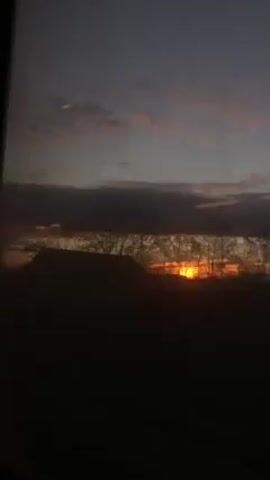 The video shows a sunrise through a window. The sky is a mix of dark blue and pink hues, with clouds obscuring most of the sun. The foreground is dark, with silhouettes of buildings and trees.
