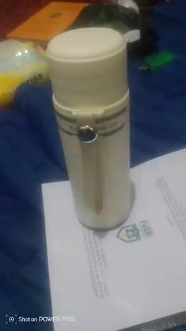The video shows a close-up, slightly blurry, vertical shot of a cream-colored thermos bottle standing on a white document. The thermos bottle has a textured pattern and a band around the middle with text that is difficult to read. There is a silver button or knob on the front of the thermos. The document beneath it has text and a logo that includes the letters "IBI". The background is a dark blue fabric, possibly a bedspread, with some out-of-focus objects including what appears to be a green lighter and some crumpled paper. The bottom left corner of the video displays "Shot on POWER PG5". The overall lighting is dim.