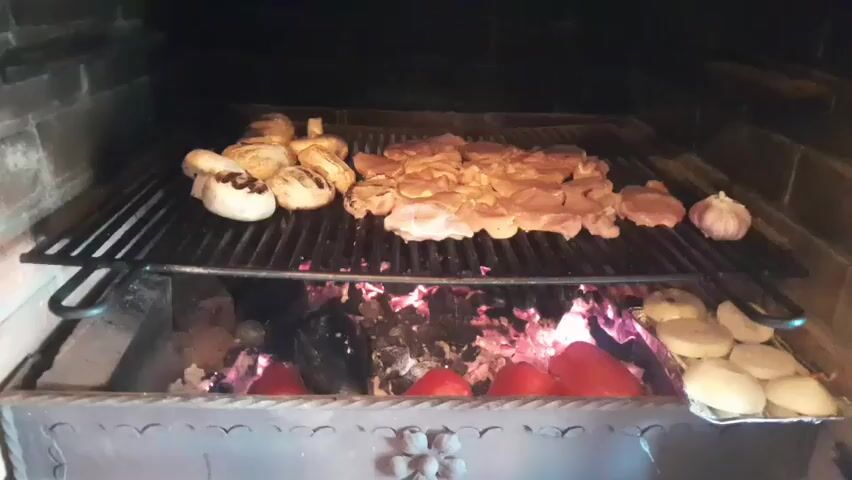 The image depicts a close-up view of food being grilled over hot coals in an outdoor barbecue. The main subjects are the food items: several pieces of chicken and some large mushrooms are arranged on a metal grill rack. Below the grill, bright red bell peppers and what appear to be potatoes are being cooked directly on the embers or on a separate tray. The setting is an open brick barbecue pit, with glowing coals visible beneath the grill. The background is dark, suggesting it might be evening or the area is dimly lit. The scene evokes a sense of casual outdoor dining or a culinary preparation for an event. There is no visible text in the image. The location context provided indicates Gliczarów Górny, Poland, which aligns with a rural or suburban outdoor setting. The lighting and the presence of a barbecue suggest a pleasant weather day, likely warm enough for outdoor cooking.