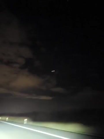 A video records a nighttime scene from a moving vehicle. The perspective is from the driver's seat, looking forward and slightly upward. A dark, cloudy sky dominates the upper portion of the frame, with faint stars visible. In the center of the sky, there appears to be a bright, unidentified object or objects, possibly a cluster of lights, that remain relatively stationary throughout the short clip. The lower half of the frame shows a dark road with a white line marking the lane. The headlights of the vehicle illuminate the road ahead. Orange and white markers are visible along the side of the road, indicating the edge of the roadway. The video captures a brief period of time, showing minimal changes in the scene.
