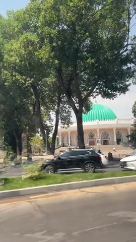 The video shows a bright, sunny day with lush green trees. In the background, a white building with a large, vibrant green dome is visible. The building appears to be an important structure, possibly a mosque or a religious center, given the dome's design. In the foreground, a street with several cars driving by is depicted. The cars are of various colors and models, indicating a typical urban environment. The overall scene suggests a moment captured while traveling, perhaps from a moving vehicle, offering a glimpse of a city's architecture and daily life.