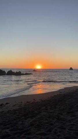 The video shows a sunset over the ocean. The sun is low on the horizon, casting an orange glow across the sky and water. Waves gently lap at the shore, which is covered in dark sand. Rocks jut out of the water on the left side of the frame, and a single rock formation is visible in the distance on the right. The sky is a clear gradient of blue and orange.