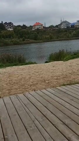 The video opens on a sandy beach next to a body of water. A wooden walkway runs along the foreground. Buildings are visible on a hill across the water. The camera then pans to the right, showing the wooden walkway continuing alongside a paved path. There are trees and some modern wooden structures. The camera continues to pan right, showing a street lamp and trees. The scene then shifts back to the left, showing more of the lake and beach. Two people are sitting on the sand near a tree. The camera then pans slightly to the right, showing the wooden walkway extending towards the lake.