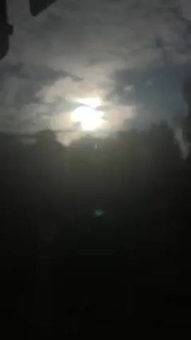 The video shows a view from inside a vehicle, looking out through the windshield at a partly cloudy sky with the sun shining through the clouds. There are trees and a tall building in the background, and some cars and a white trailer visible below. The camera appears to be moving, possibly as the vehicle is driving.