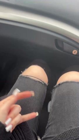 Here's a description of the video:

The video starts with a view from the passenger seat of a car, showing a person's legs clad in ripped black jeans. The person's hand is visible, making a peace sign. 

The view then shifts to the car's dashboard and the windshield. The dashboard features a built-in display showing information such as time and radio frequency. Raindrops are visible on the windshield, and the outside view reveals a cloudy day with a green landscape, potentially a field or grassy area, and what seems to be a mountain or hill in the distance. The car is moving.