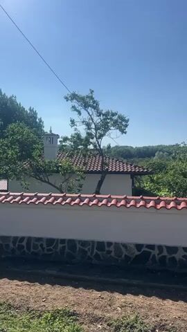 A video pans across a whitewashed wall topped with red tile, a stone foundation, and green grass. Beyond the wall, a white building with a red-tiled roof is visible, with a tree growing in front of it. Further in the background, lush green trees and foliage cover rolling hills under a clear blue sky. The sun shines brightly, creating lens flare in the upper right corner of the frame. A thin black wire stretches diagonally across the sky.