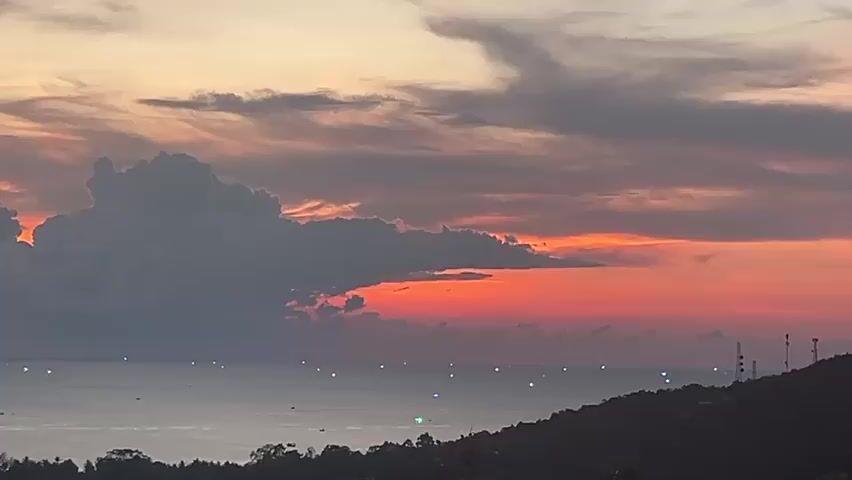 The video shows a wide shot of a sunset over the ocean. A large, dark cloud hangs in the left side of the frame, partially obscuring the horizon. The sky displays a gradient of colors, ranging from orange and red near the horizon to gray and peach higher up. The sea is relatively calm with scattered points of light visible across its surface. A dark, hilly landscape with vegetation and structures, including tall antennae or towers, is silhouetted in the lower right corner of the frame.