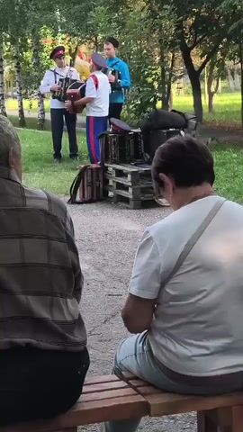 The video shows an outdoor performance in a park-like setting. Three men in traditional Cossack-style attire are playing accordions and a balalaika. They are standing on a gravel path, with a grassy area and trees behind them. In front of the musicians, two individuals, appearing to be audience members, are seated on a wooden bench, facing away from the camera. Further in the background, several women in red dresses are also present, some standing and some seated. The overall atmosphere suggests a cultural event or a folk performance.
