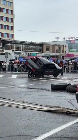 In an outdoor urban setting, under an overcast sky, a black car is overturned on its side. Several individuals in what appear to be firefighter uniforms are interacting with the vehicle, suggesting a rescue or demonstration. A crowd of spectators is visible behind a safety barrier in the background, some holding umbrellas, indicating potential rain. Several large tires are lying on the wet asphalt near the overturned car. In the foreground, a red and white vehicle, possibly a truck or van, is partially visible on the right side of the frame. The scene appears to be part of a public event or demonstration.