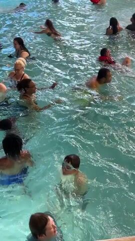 The video shows a crowded indoor swimming pool, likely a water park. Numerous people of various ages are in the water, some swimming, others wading, and some are playing. The water is a light blue and appears to be somewhat agitated, with ripples and splashes visible throughout the scene. The lighting is bright, suggesting artificial lighting typical of an indoor facility. People are wearing swimwear, and some children are wearing goggles. The overall atmosphere appears to be one of recreation and enjoyment.