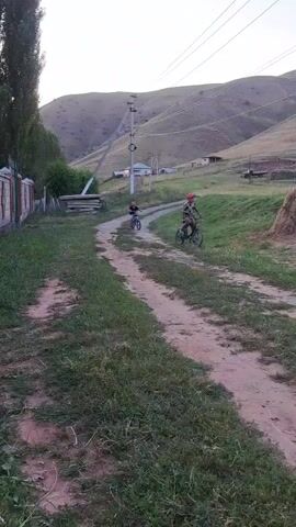 A grainy, low-resolution video shows two children on bicycles on a dirt road in a rural, hilly area. The child in the foreground is on a blue bicycle, wearing a dark t-shirt and shorts. The child in the background is on a bicycle and appears to be wearing camouflage clothing and a red helmet. They are riding away from the camera, up a slight incline. In the background, there are houses, a car, power lines, and a large haystack. The overall impression is of a casual, outdoor activity in a semi-arid landscape.