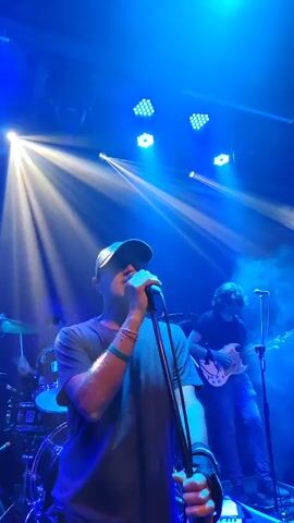 A rock band performs under bright blue stage lights and fog. The lead singer, wearing a grey shirt and baseball cap, is close to the camera, singing into a microphone. A guitarist in a dark shirt and jeans plays a white electric guitar to his right. Behind them is a bassist in a white t-shirt and jeans, also singing into a microphone. The stage is cluttered with equipment, including keyboards and drum sets. The venue appears to be a dark club or theater, with speaker stacks visible overhead.