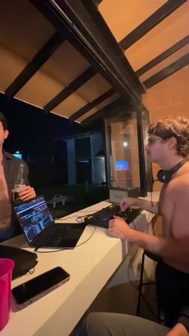 Two young men are visible outdoors at night, seemingly at a party. One man, wearing a dark shirt, stands on the left, holding a bottle of beer. The other man, shirtless and wearing glasses and headphones, is seated on the right, operating a DJ setup that includes a laptop and mixing board. A pink cup and a smartphone are on the counter in front of them. The background shows a dimly lit yard and the side of a building.