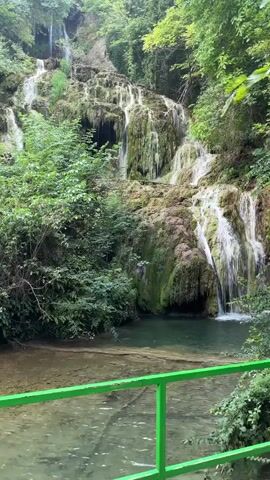 This is a short video clip of a waterfall in a lush, green forest. The waterfall cascades down a rocky cliff face, with water flowing over moss-covered rocks and vegetation. The water collects in a clear pool at the bottom of the falls, where it then flows away. A bright green metal railing is visible in the foreground, suggesting that this is a scenic overlook or a designated viewing area for visitors. The overall atmosphere is serene and natural, with the sound of the waterfall being the dominant audio element.