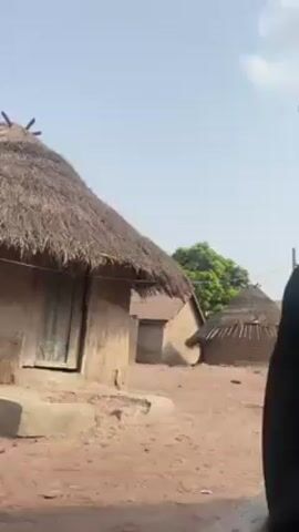 The video shows a scene in what appears to be a rural village. The camera is initially pointed upwards towards the sky and then pans down to show two men sitting under a thatched roof structure. The men appear to be resting. The background shows several huts with thatched roofs, typical of rural architecture. One man is wearing a black shirt and appears to be looking upwards, while the other is wearing a cap.  The video ends with a close-up of a wooden surface and a pair of black sandals underneath it.