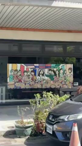 The video shows a colorful painting of the Last Supper on the window of a building. A car is parked in front of the building. Potted plants sit on the sidewalk.