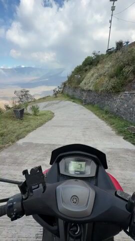 The video captures a first-person perspective of riding a red Yamaha scooter on a winding concrete road. The rider is navigating a downhill path with a stunning vista of mountains and a valley spread out before them. The sky is a vibrant blue, dotted with fluffy white clouds, suggesting a clear day with perhaps some cloud cover at higher elevations. The road itself is flanked by lush green grass and a stone wall covered in small white flowers on the right side.

In the distance, a vast expanse of land, possibly a caldera or a large plain, is visible, with mountains forming the backdrop. There are trees scattered throughout the landscape, and a small building with a red roof is situated to the left of the road in the middle distance. A utility pole stands tall on the right side of the road, near the crest of the hill.

The scooter's digital display is visible, showing a fluctuating speed, ranging from 10 to 16 km/h, indicating a leisurely pace, likely for sightseeing. The overall mood is serene and adventurous, with the beautiful natural scenery creating a sense of peace and wonder. The location is identified as Cemorolawang, Indonesia, known for its proximity to Mount Bromo, which the expansive landscape might suggest. The time of day appears to be morning or early afternoon, given the brightness of the sunlight and the clarity of the sky. There is no visible text other than the "YAMAHA" logo on the scooter's dashboard.
