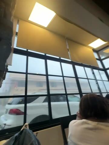 The video shows a view through a window with raindrops on it. Outside, a white car is parked in front of a building. A person's back is visible in the foreground, facing away from the camera and towards the window. The weather appears to be overcast or raining.