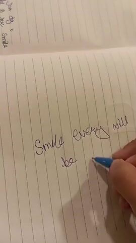 A person's hand is holding a blue pen and writing in a lined notebook. The pen is writing the words "Smile every will be fine" on the page. The notebook is open, and the pages are lined. There is some text written on the top left of the page, but it is mostly out of focus. The lighting suggests an indoor environment.