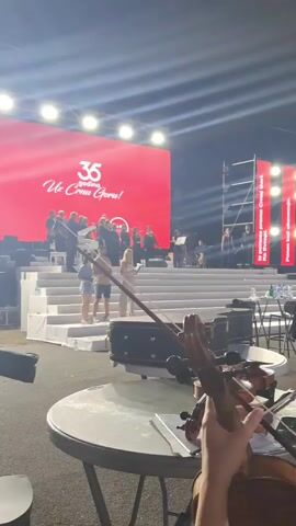 The video shows an outdoor stage at night. A large LED screen behind the stage displays "35 Uzdina" and other text in red and white. A choir of approximately 15 people stands on a tiered platform in front of the screen, some holding sheet music and standing behind music stands. Two people in casual clothing are visible on the lower steps of the platform. To the right of the stage, scaffolding and vertical banners are present. In the foreground, a person's hands hold a violin and bow, with sheet music on a table in front of them. The area in front of the stage appears to be a performance space or backstage area.