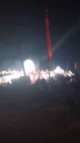 The video shows a night scene at what appears to be an outdoor event or carnival. In the foreground, a dark, uneven surface suggests dirt or rough ground. To the left, there are silhouetted figures of people and possibly motorcycles. A red and yellow flag is visible on a pole to the right. In the background, a large, brightly lit Ferris wheel is the central attraction, with tents and other structures illuminated around it. People are gathered in front of the tents, and there are lights illuminating the area, creating a festive atmosphere. The sky is dark, indicating it is nighttime. The video is shaky, suggesting it was filmed handheld.