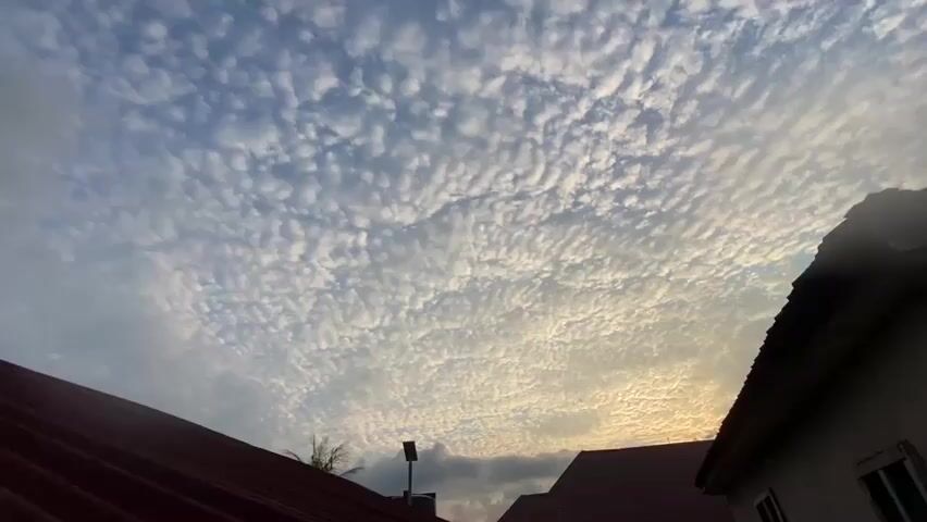 This media file captures a wide-angle view of a sky filled with altocumulus clouds, creating a textured, ripple-like pattern. The clouds are predominantly white and gray, with patches of golden light suggesting the sun is low on the horizon, indicating either dawn or dusk.

In the foreground, the roofs of two houses are visible, one with a dark brown corrugated metal roof on the left, and another with a reddish-brown tiled roof on the right. The house on the right also shows a section of its white exterior wall and a window. A street lamp stands tall against the sky, positioned between the two houses, suggesting a residential or urban setting. A silhouetted tree is visible to the left of the lamp post.

The overall scene is peaceful and serene, dominated by the striking cloud formation and the warm glow of the setting or rising sun. There are no people or animals visible in the frame. The location context provided is Igbara, Nigeria, which aligns with the typical architecture and weather patterns for such a region. The time of day is clearly indicated by the quality of light and the cloud types, pointing towards either early morning or late evening. There is no visible text in the media file.